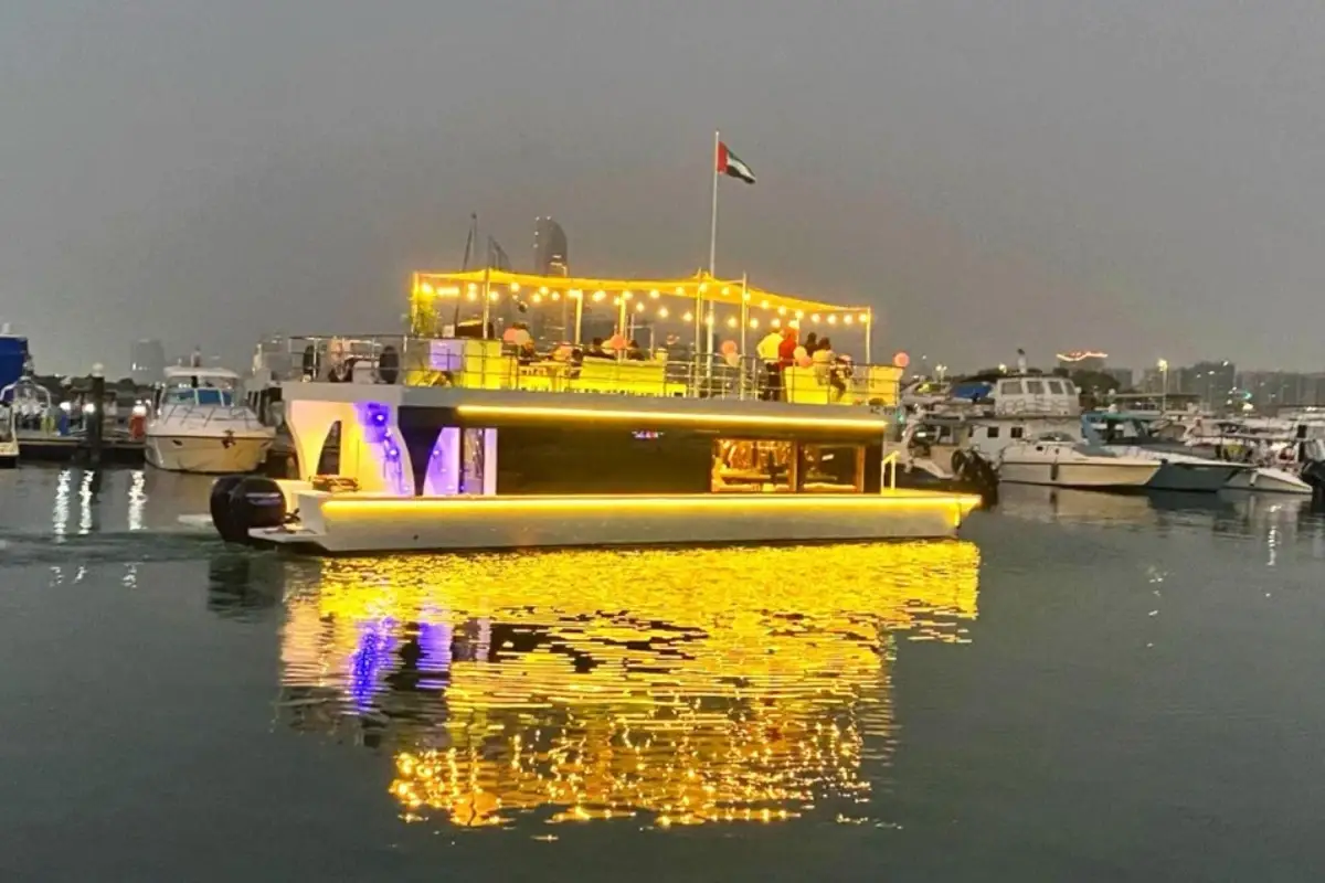 Haya House Boat at dusk at the marina, its upper deck strung with lights and guests on top, the hull lit in yellow and reflected in the water with the Abu Dhabi skyline behind